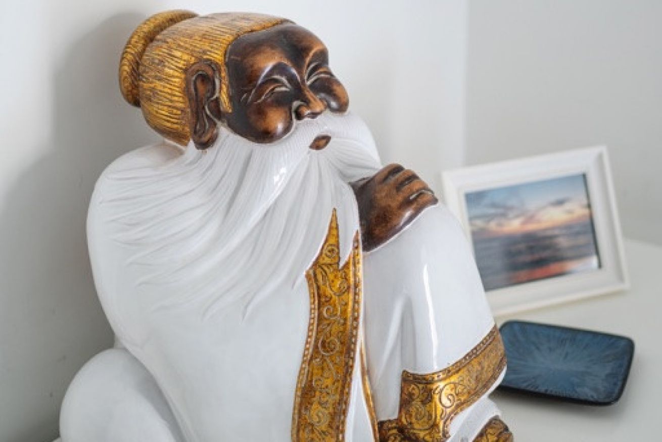 Eine Statue einer in einen weißen Gewand mit goldener Bordüre gekleideten Gestalt, vor einer schlichten Wand mit einem gerahmten Bild und einem blauen Gegenstand in der Nähe.