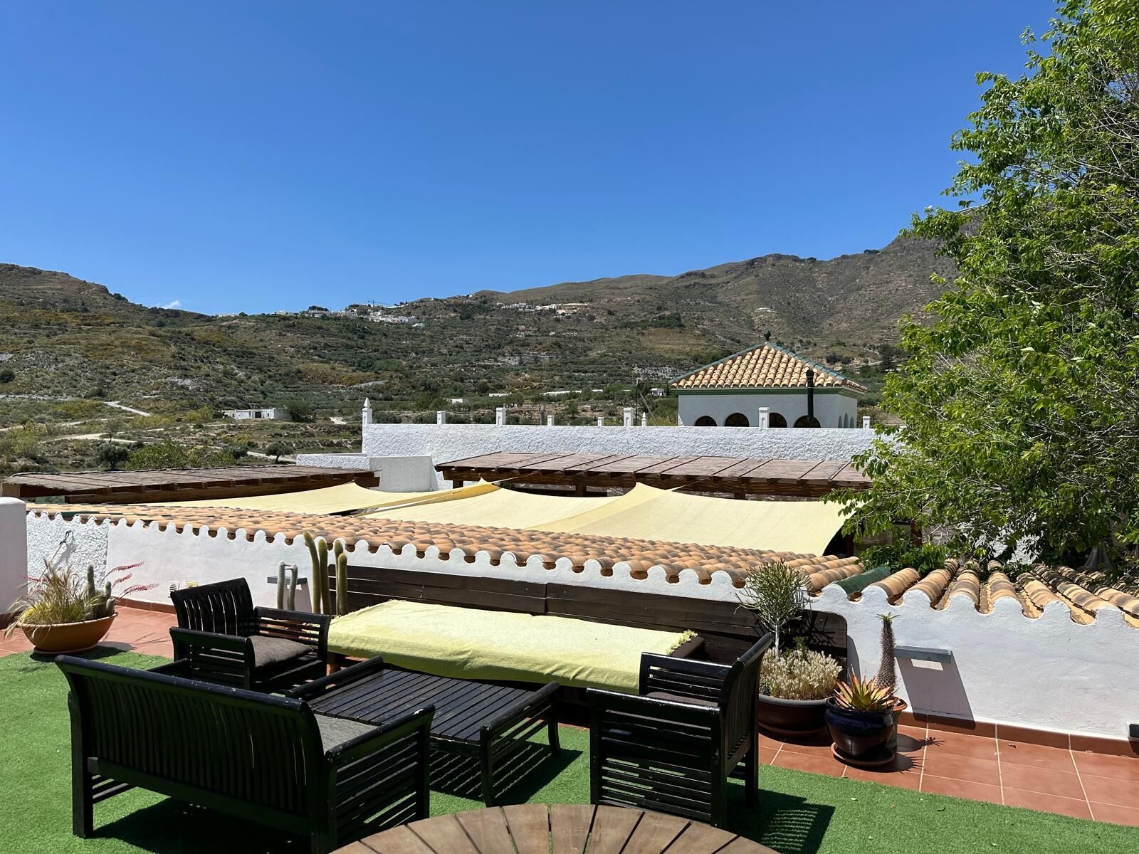 A rooftop terrace with outdoor furniture, a view of a mountainous landscape, and a building with a tiled roof in the background.