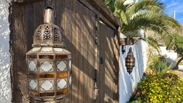 A traditional lantern attached to a wooden door, with a palm tree and a wall with yellow flowers in the background. - ALQUERIA DE GÍTAR. Beautiful rural villa for 20 people close to the sea (Felix)