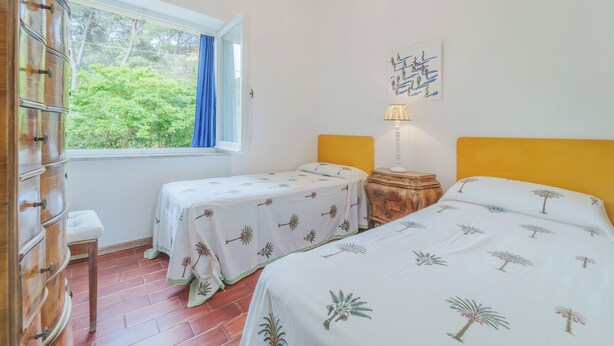 A room with two beds, a wooden dresser, a window with a view of greenery, and a wall-mounted lamp. - A Country House by the Sea (Procchio)