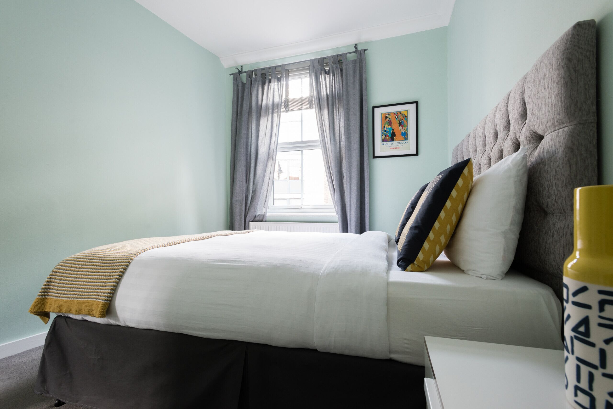 A bedroom with a bed, a window with curtains, a framed picture on the wall, and a bedside table with a lamp.