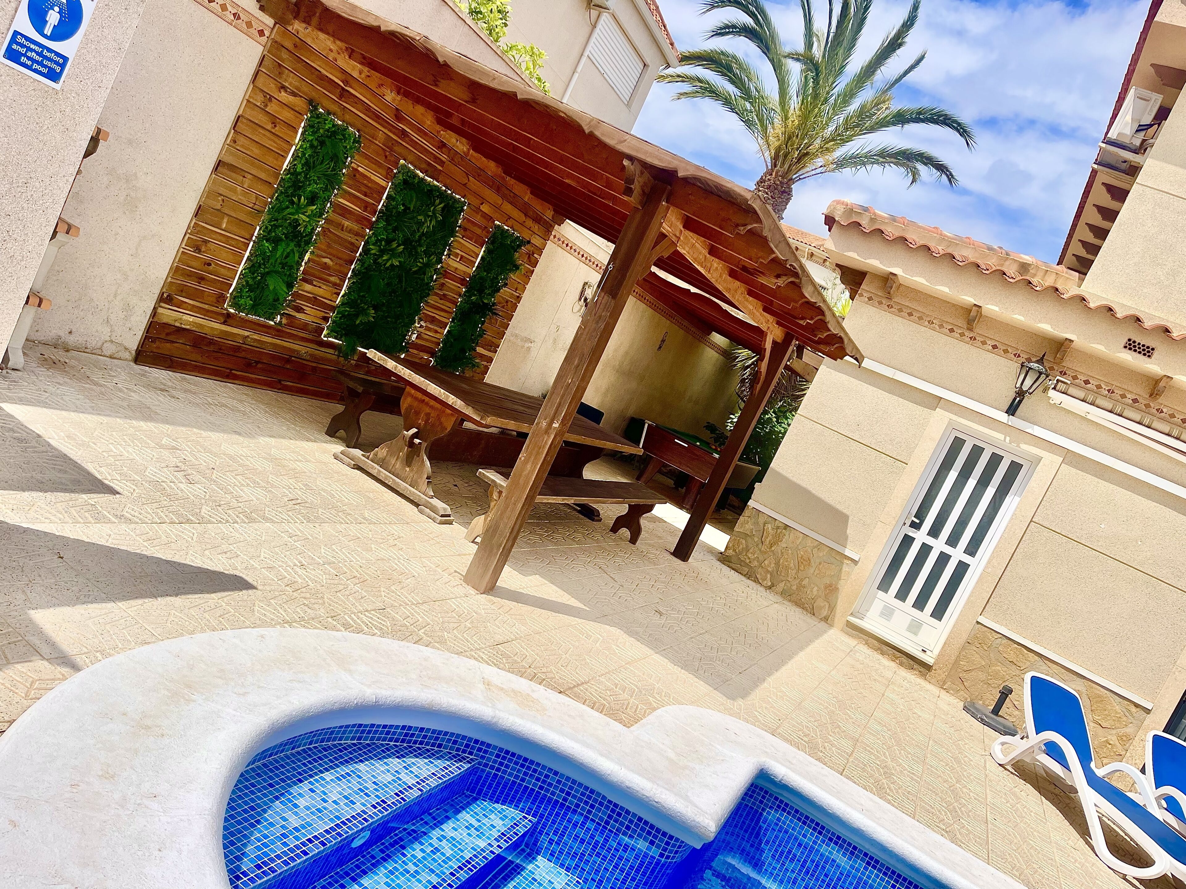 Un área de piscina con una pérgola de madera, una palmera y suelo de baldosas.