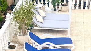 Un área de estar al aire libre con luz solar, una pérgola blanca, sillones azules y diversas plantas en macetas.