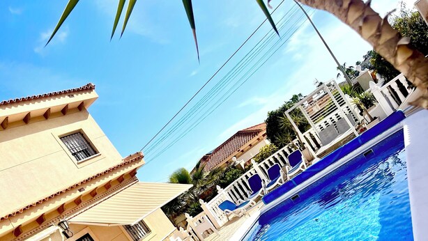 Una piscina con borde de baldosas, cerca blanca y un edificio con techo y ventanas de cerámica.