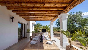 Un espace extérieur aménagé pour les repas, avec une pergola en bois, des colonnes blanches et des plantes en pot.