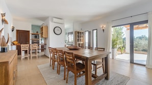 Un vaste espace de salle à manger avec une table et des chaises en bois, une cuisine en arrière-plan et une grande porte-fenêtre coulissante donnant sur un espace extérieur.