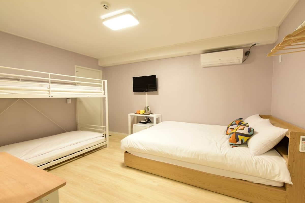 A room with a bunk bed, a single bed, a desk, a TV, and an air conditioning unit.