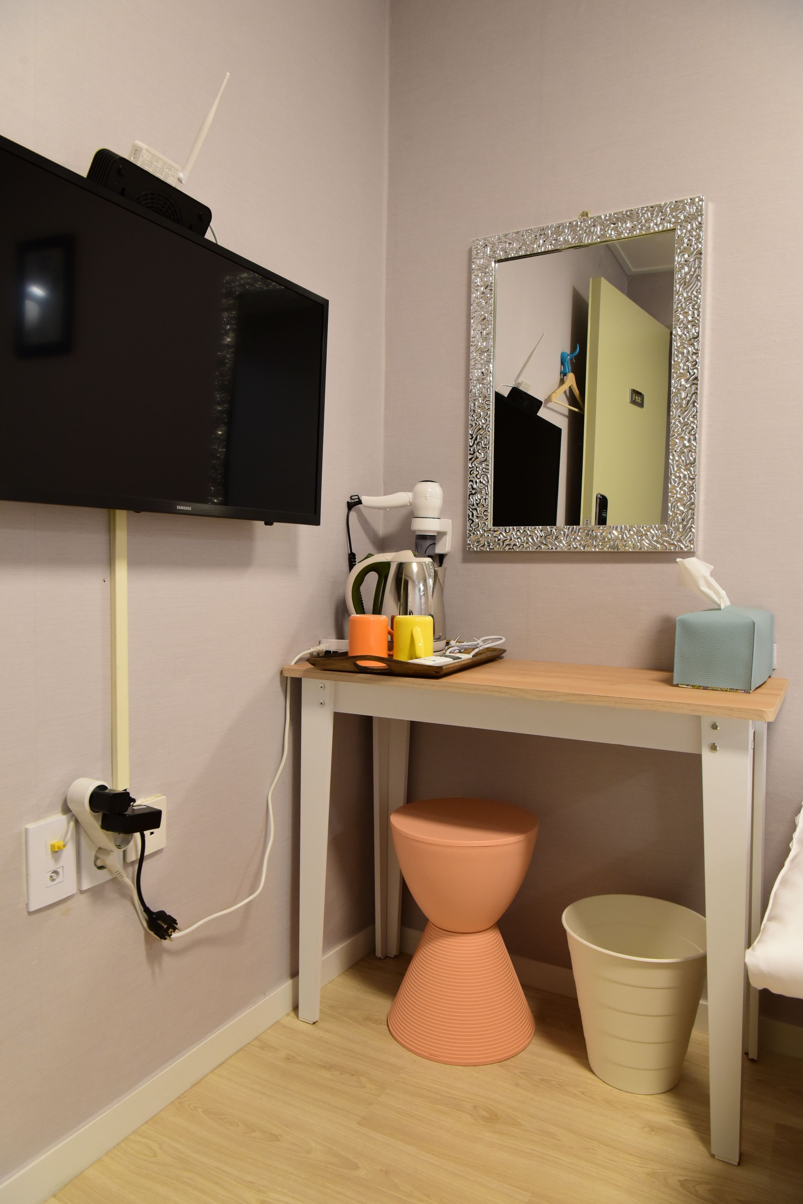 A small room with a TV mounted on the wall, a mirror with a decorative frame, a wooden desk with a tissue box, a trash bin, and a small orange stool.