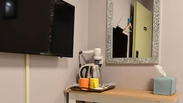 A small room with a TV mounted on the wall, a mirror with a decorative frame, a wooden desk with a tissue box, a trash bin, and a small orange stool.