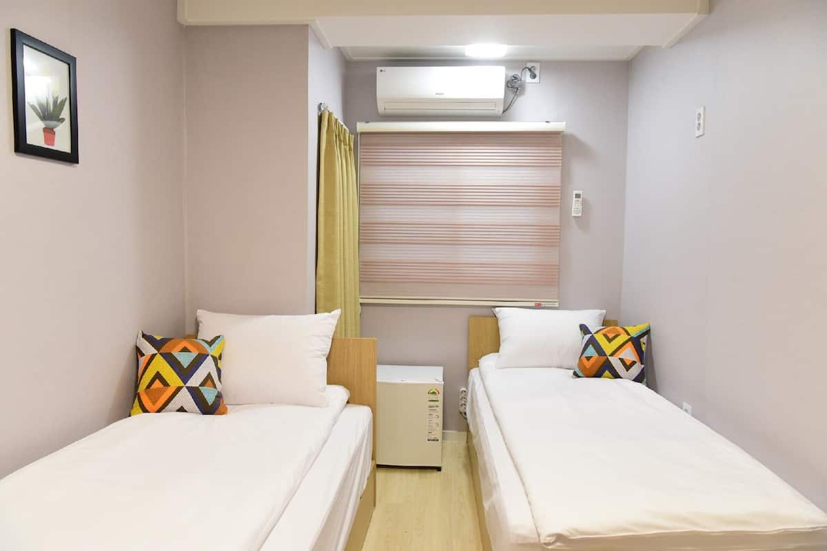A small, well-lit room with two beds, a small air conditioning unit, and a window with blinds.