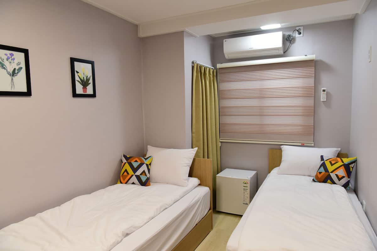 A hotel room with two single beds, a small bedside table, a wall-mounted air conditioner, and a window with blinds.