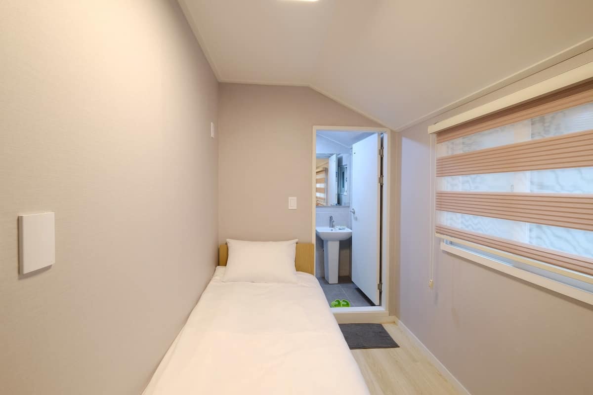 A narrow hotel room with a single bed, a small bathroom, and a window with blinds.