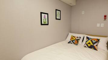 A neatly made bed with white bedding and two colorful geometric pillows, a plain wall with two framed botanical prints, and a visible fire alarm.