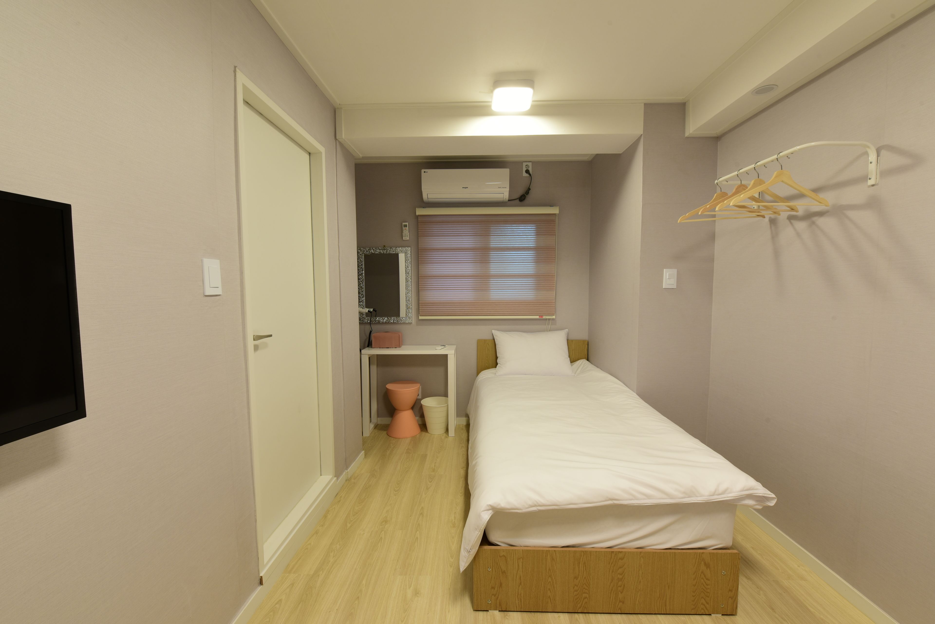 A small, neatly arranged bedroom with a single bed, a bedside table, a hanging clothes rack, and a wall-mounted air conditioner.