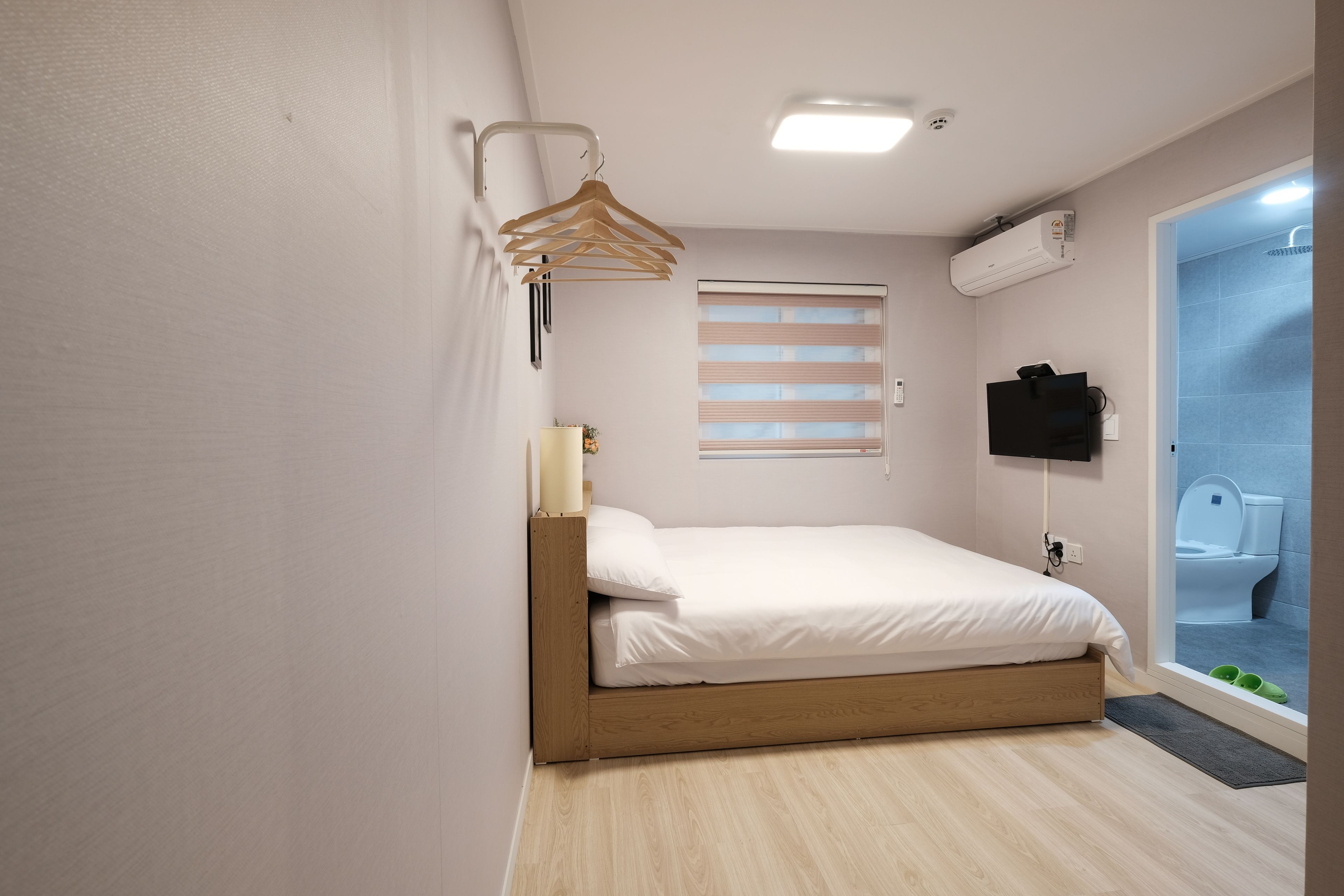 A small, modern bedroom with a single bed, a wall-mounted TV, a wooden hanger, and a window with blinds.