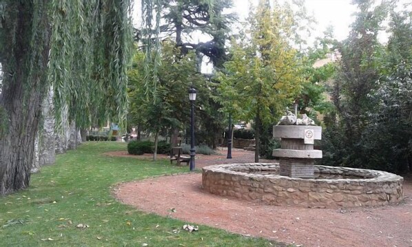 Une fontaine en pierre ornée d’une sculpture d’oiseau, entourée d’une bordure en pierre, dans un parc arboré avec un banc.