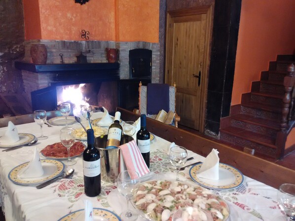 Una mesa de comedor con platos, cubiertos, botellas de vino y copas, con una chimenea y una escalera al fondo.