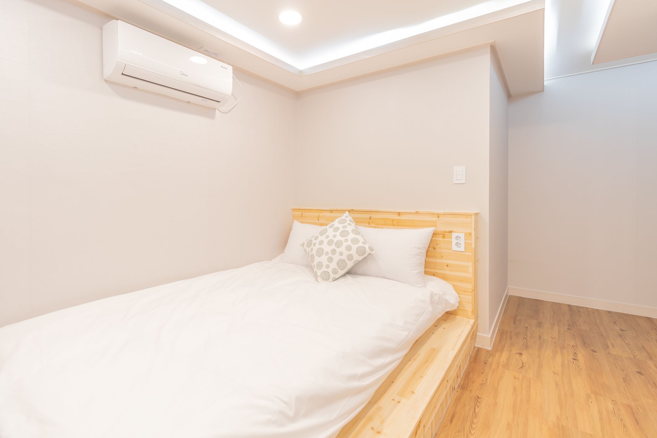 A single bed with a wooden headboard, white bedding, and a polka dot pillow. An air conditioning unit is mounted on the wall above the bed.
