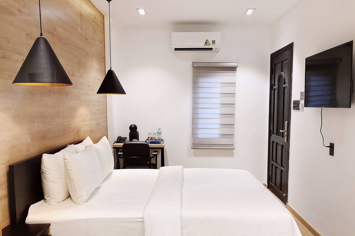 Une chambre d’hôtel comprenant un lit, deux lampes suspendues noires, un bureau avec une chaise, un téléviseur fixé au mur et un appareil de climatisation.