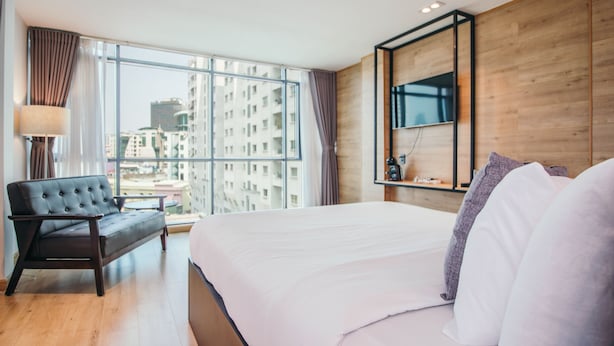 Una habitación de hotel moderna con una cama grande, un sofá, un televisor montado en la pared y vistas de la ciudad a través de amplias ventanas.
