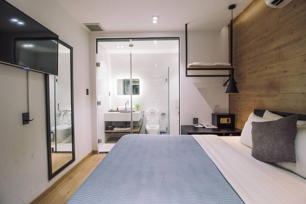 Une chambre d’hôtel moderne avec un grand lit, une tête de lit en bois, une table de chevet avec une lampe, une armoire et une salle de bain visible à travers une porte en verre.