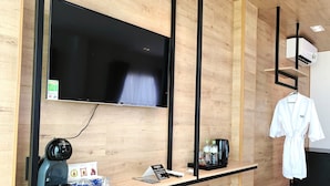 Habitación de hotel con televisor de pantalla plana montado en la pared, cafetera, un pequeño estante con artículos de aseo y una bata colgada en un perchero.