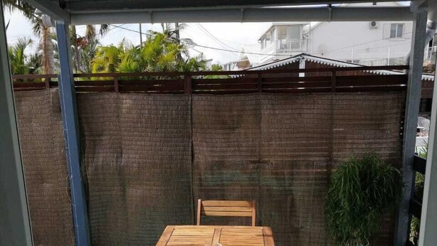 Un patio couvert avec une table en bois et deux chaises, un tapis bleu et une clôture en bambou en arrière-plan.