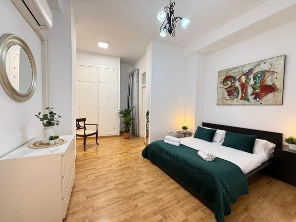 A modern bedroom with a wooden floor, a bed with green bedding, a bedside table with a lamp, a mirror, and a painting on the wall.