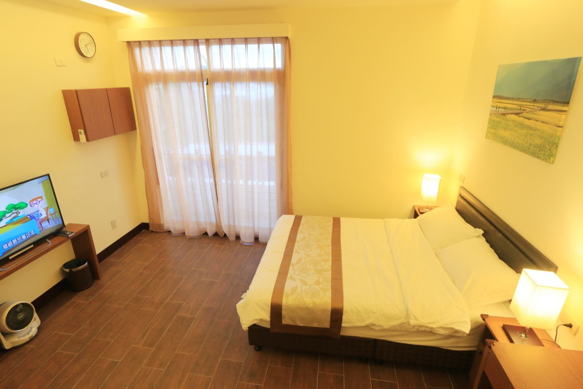 A hotel room with a bed, a TV, a clock, curtains, a painting, and a fan.