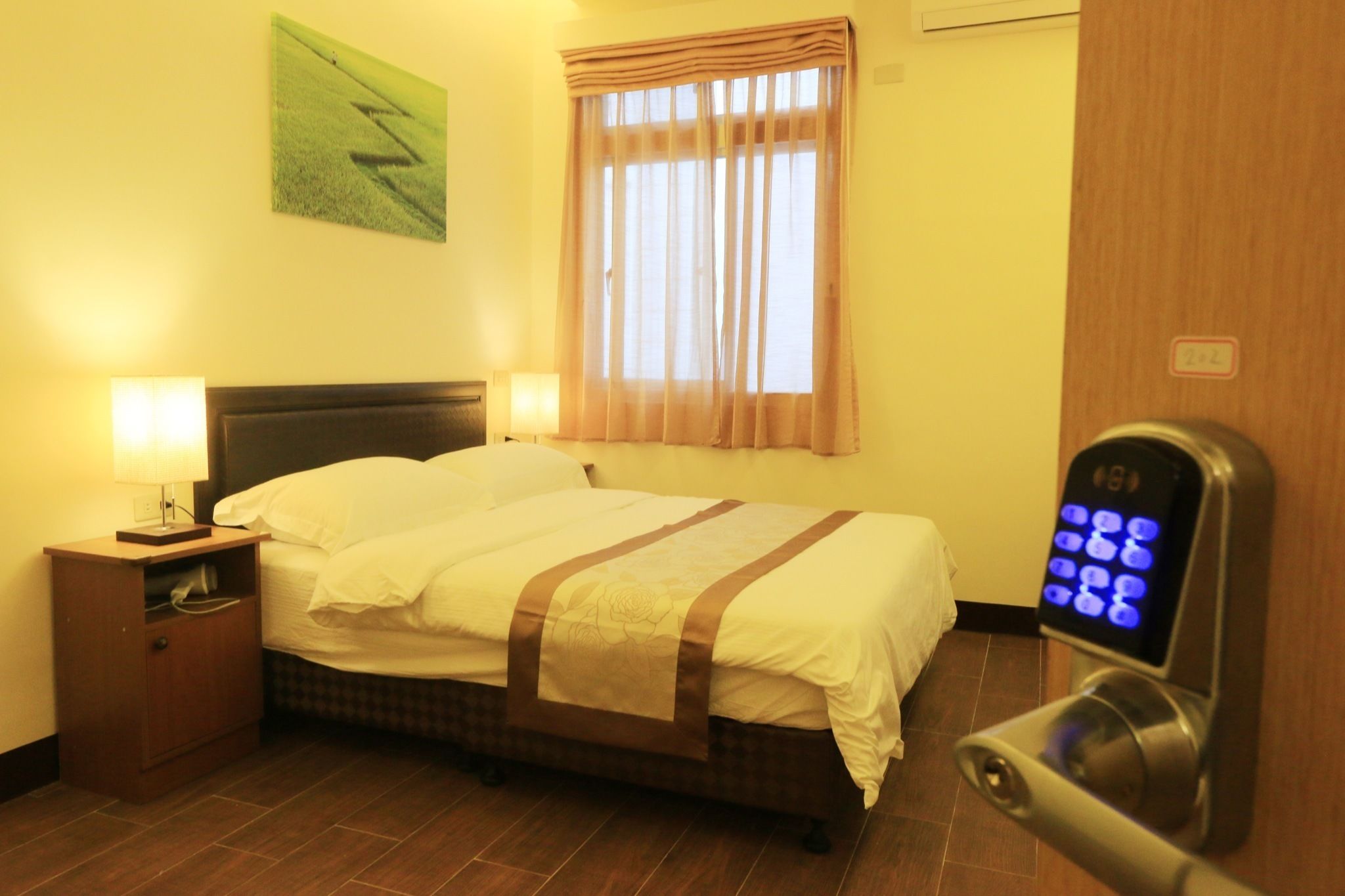A hotel room with a bed, bedside lamps, a nightstand, a window with curtains, and a digital lock on the door.