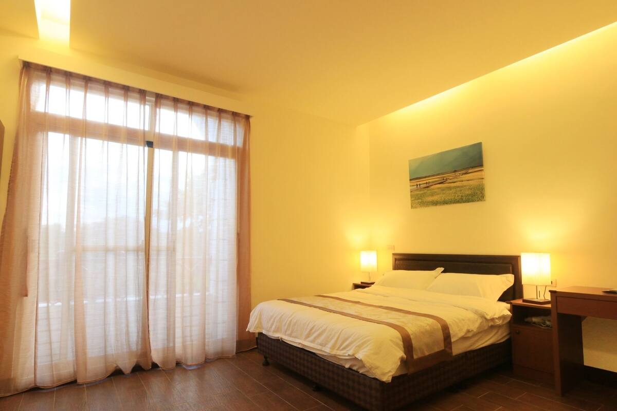 A hotel room with a large bed, two bedside lamps, a nightstand, a desk, and a painting on the wall.