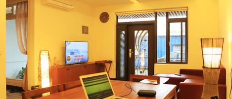 A room with a wooden table, a laptop, a television, and a wicker lamp.