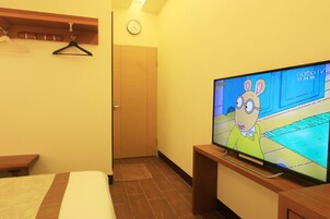 Una habitación de hotel con una cama, un televisor mostrando un dibujo animado, una puerta de madera y un reloj fijado a la pared.
