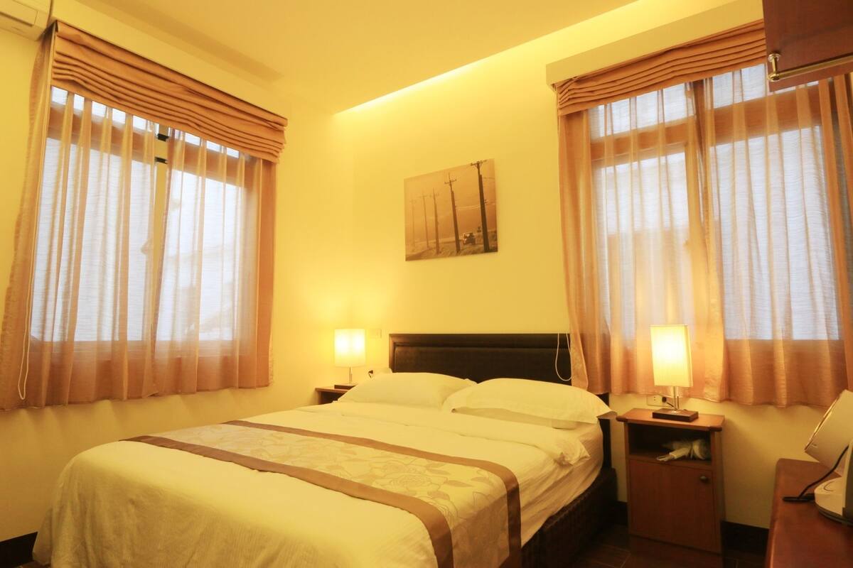 A hotel room with a bed, two bedside lamps, a nightstand, a desk with a lamp, and a painting on the wall.