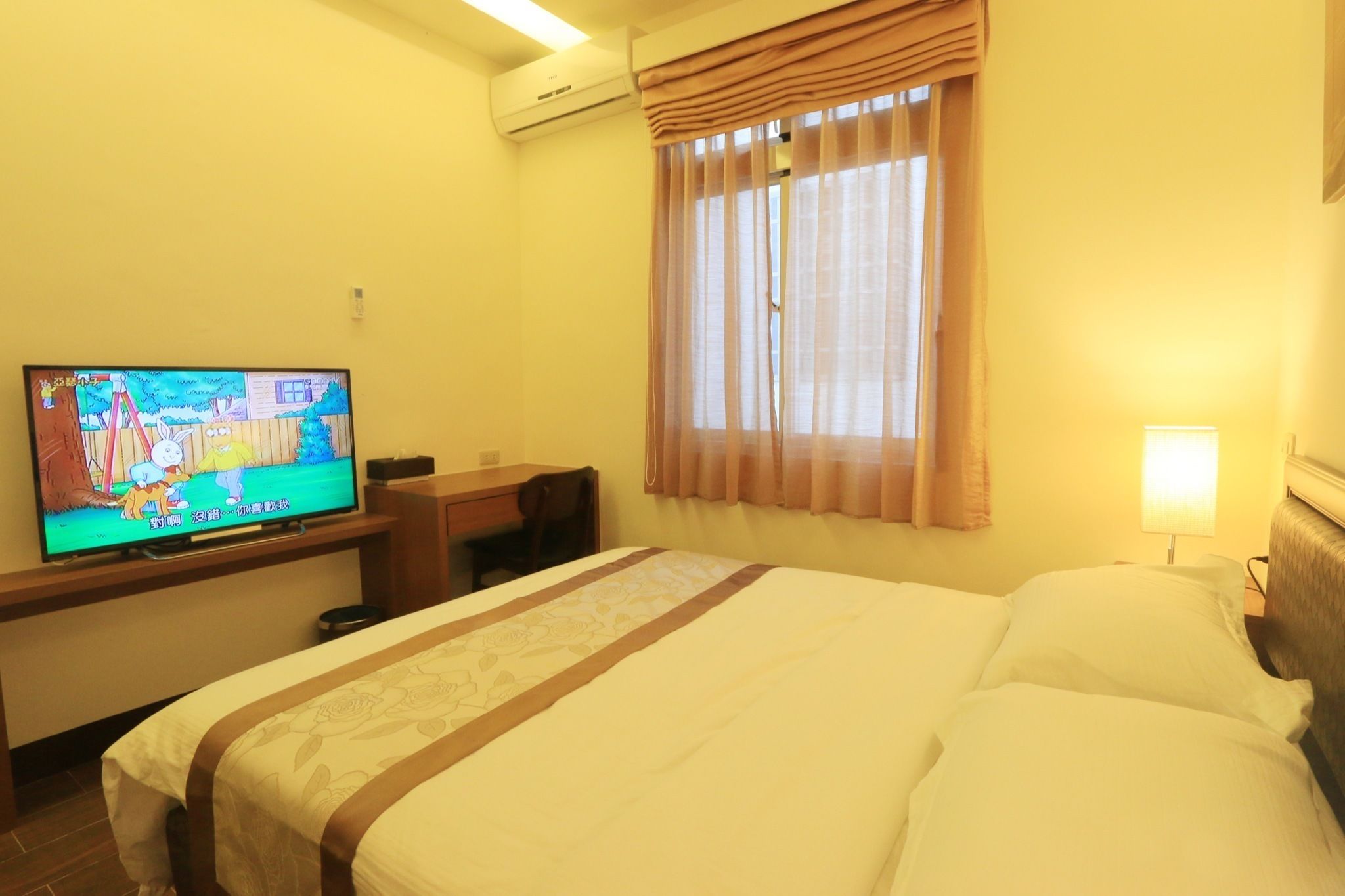 Una habitación de hotel con cama, escritorio con televisor, ventana con cortinas y lámpara.