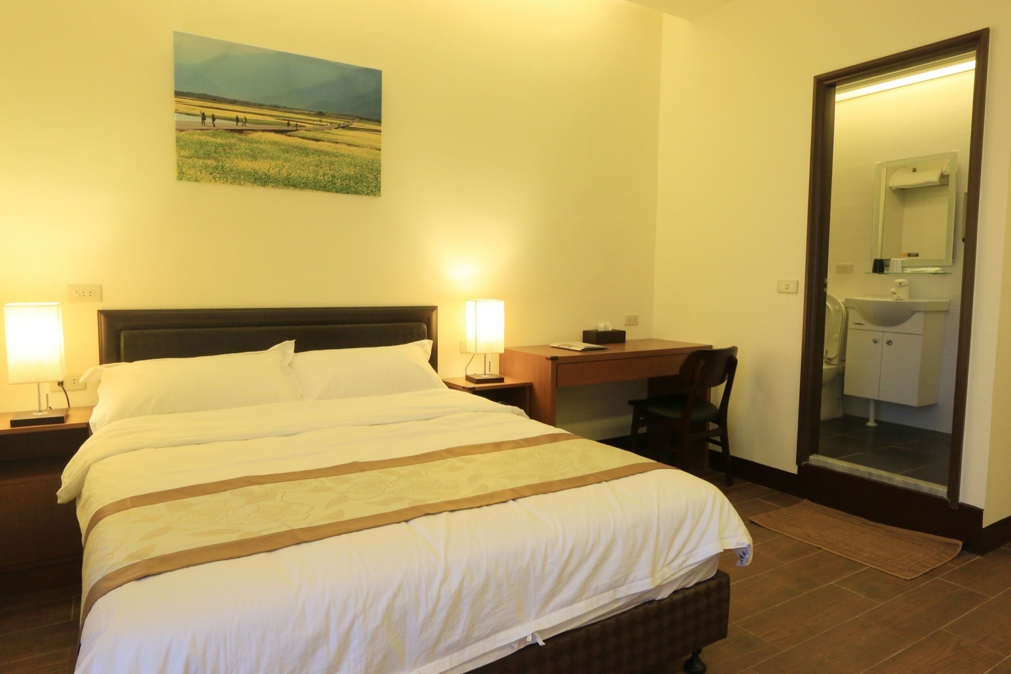 A hotel room with a large bed, a desk, a chair, a mirror, and a bathroom visible through an open door.