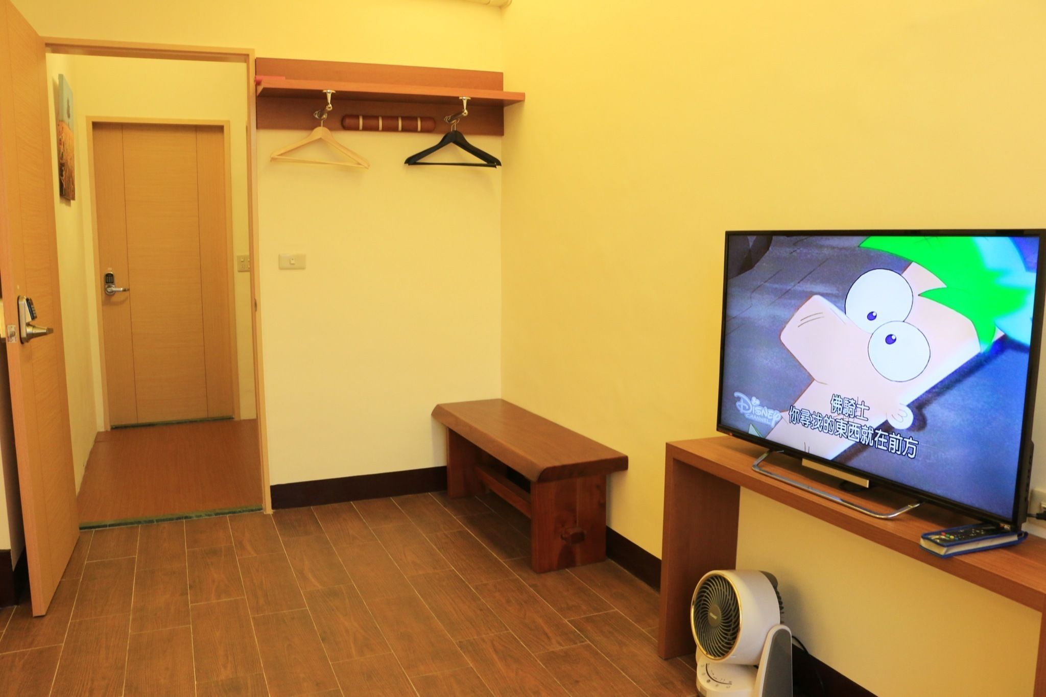 Una habitación con puerta de madera, un banco, un televisor que muestra un personaje animado y un estante pequeño con un control remoto.