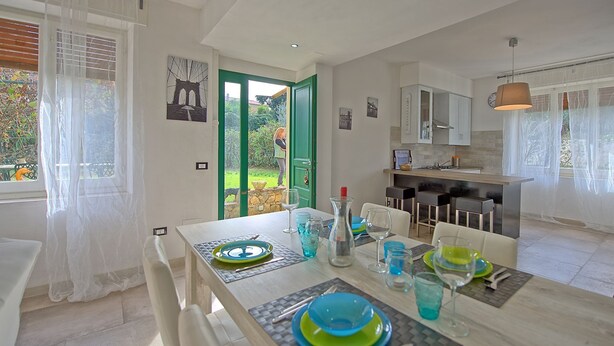 Una cocina moderna con armarios blancos, un comedor con mesa dispuesta para cuatro personas y vista al jardín a través de una puerta abierta.