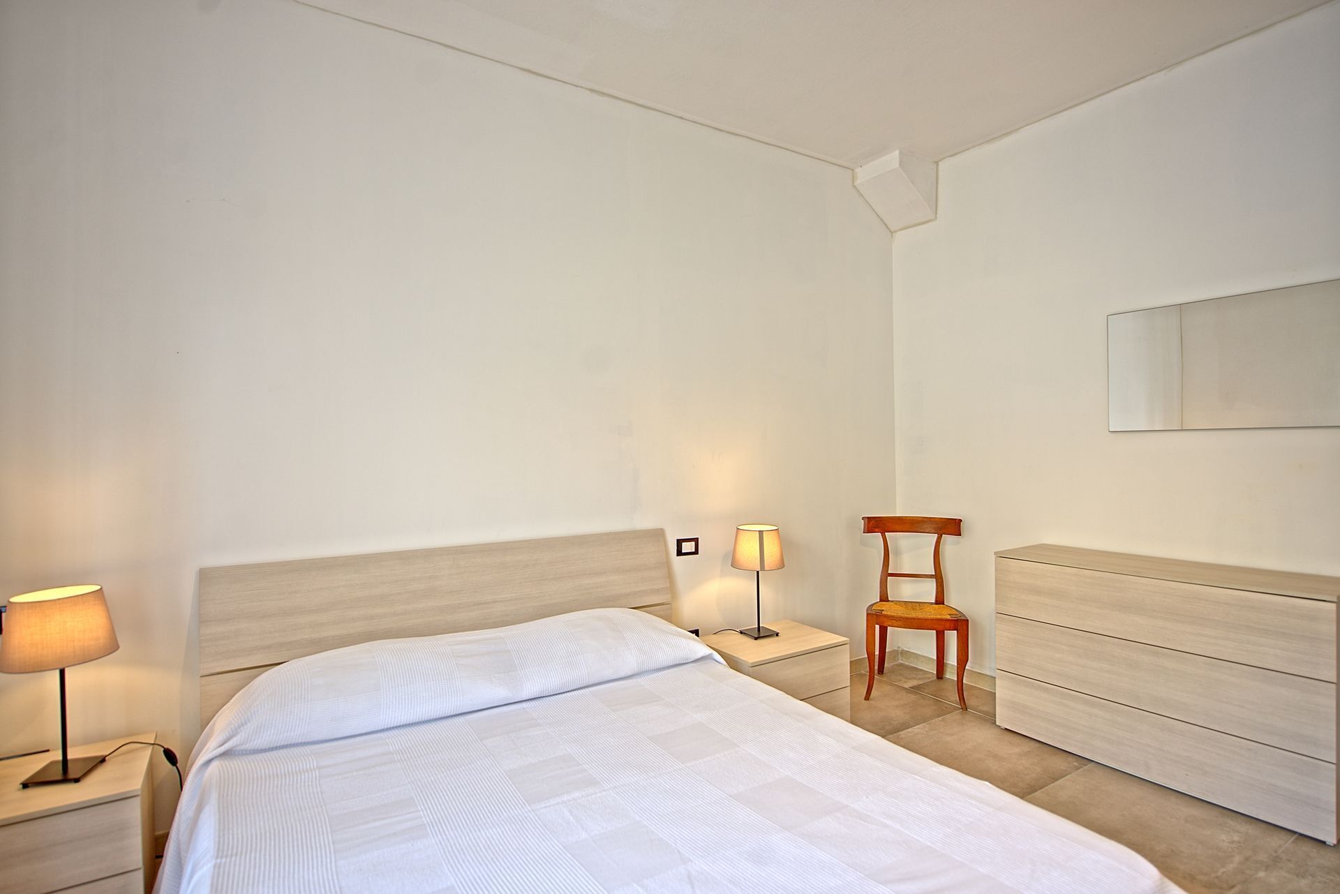 Een slaapkamer met een bed, twee nachtkastjes, een stoel, een lamp en een spiegel aan de muur.