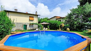 Una piscina con cubierta de madera, rodeada de abundante vegetación y dos casas al fondo.