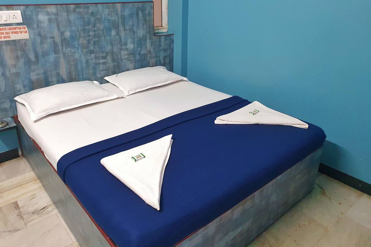 Una cama con sábanas blancas y una colcha azul, un almohadón blanco y una pared azul con una ventana.
