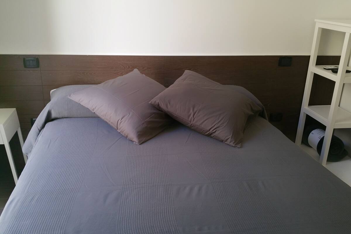 A neatly made bed with a headboard, a small bedside table with a plant, and a white shelving unit to the right.