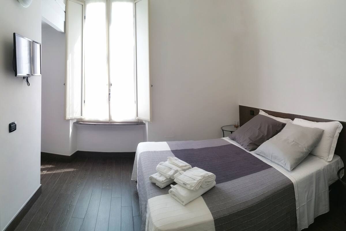 A hotel room with a bed, a window, a flat-screen TV, and a bedside table.