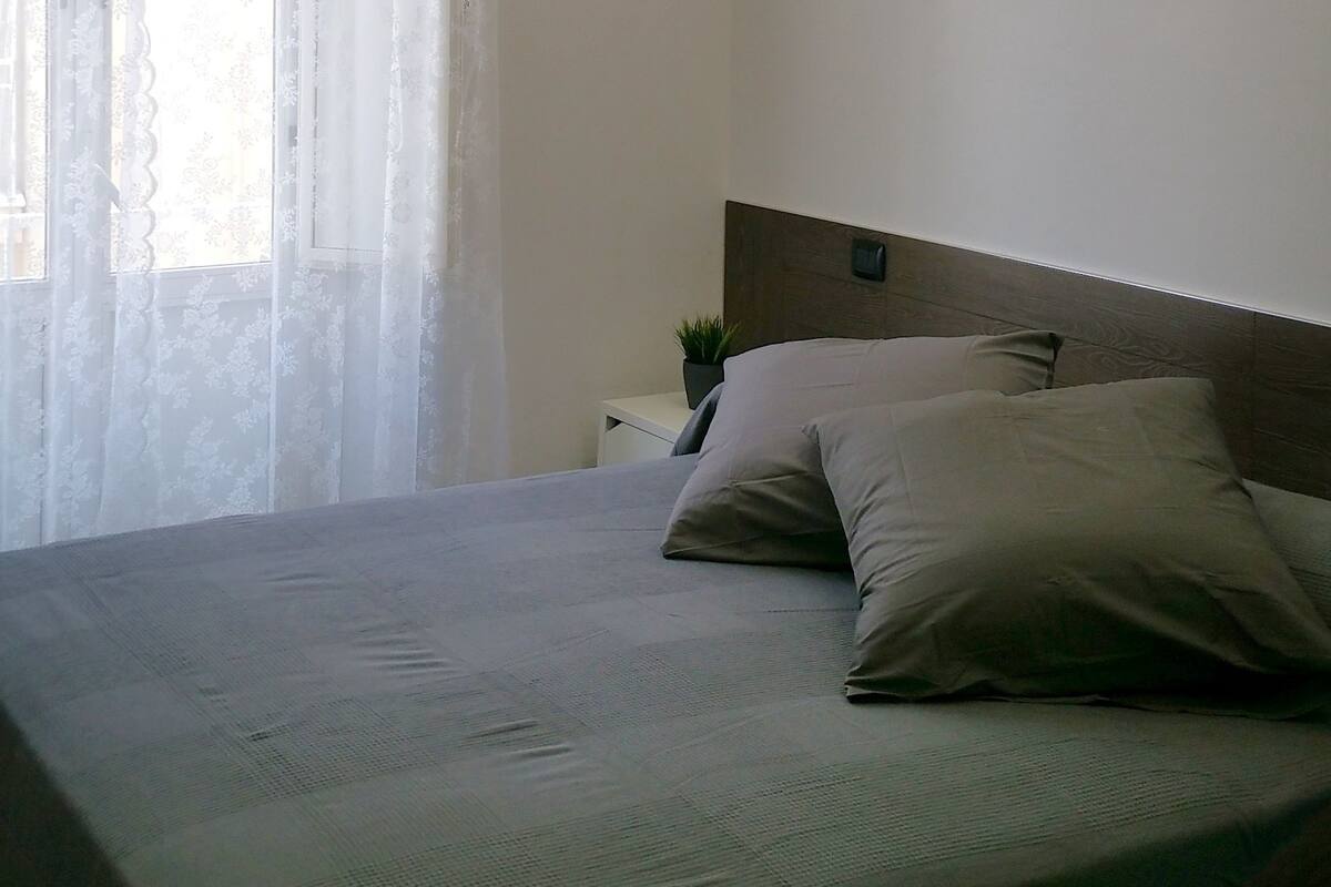 A modern bedroom with a bed, a nightstand, a potted plant, and a window with sheer curtains.