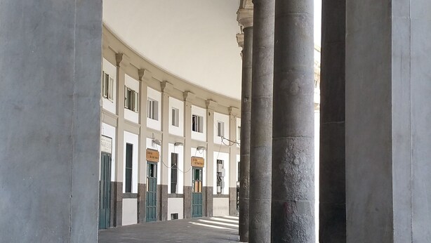 Une cour intérieure ornée de colonnes classiques et dotée d’un sol carrelé, qui mène à un bâtiment comportant de nombreuses portes et fenêtres.