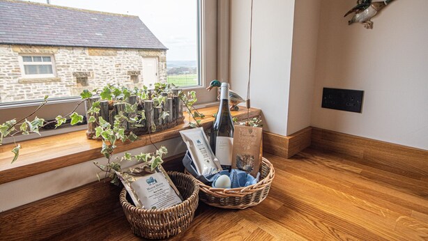 A cozy room with a large window, a stone building outside, a wicker basket with a bottle, and a decorative bird on the wall. - The Old Farmhouse - 2 Bedroom property, stunning character, views & location (Derbyshire)