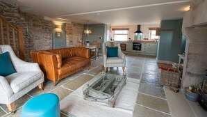 A spacious living room with a stone wall, a leather sofa, a white armchair, and a dining area with a table and chairs. - The Old Farmhouse - 2 Bedroom property, stunning character, views & location (Derbyshire)