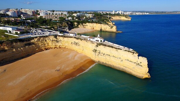 Eine Luftaufnahme einer Küstenlandschaft mit Sandstrand, einem weißen Gebäude mit spitzer Dachform und einer felsigen Klippe.