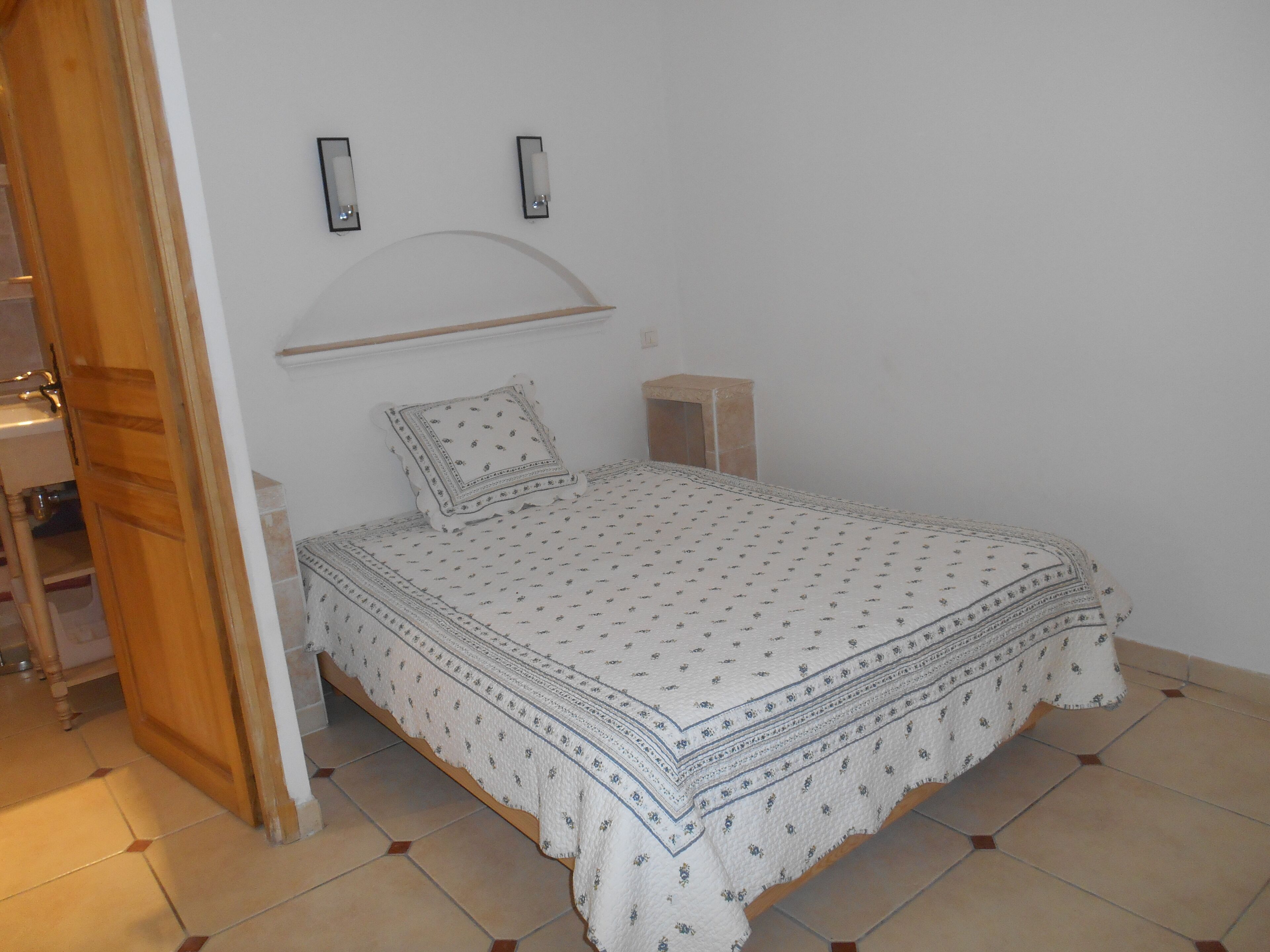 A bedroom with a bed, a wooden door, a small bedside table, and a wall-mounted shelf.