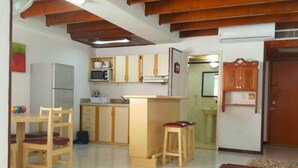 A compact kitchen with wooden cabinets, a refrigerator, and a microwave. There is a dining area with a table and chairs, and a small stool. The room has a tiled floor and a wooden ceiling. - Apartamentos 3-54 (San Andrés)